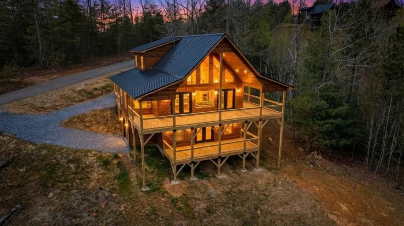 Aerial view at dusk of a wood-sided home with lit windows and a raised deck, with a curving gravel drive