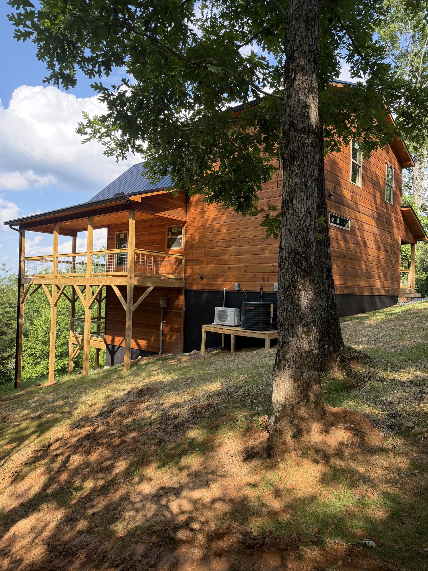 Stained cabin with covered porch on a sloped lot beside a large tree