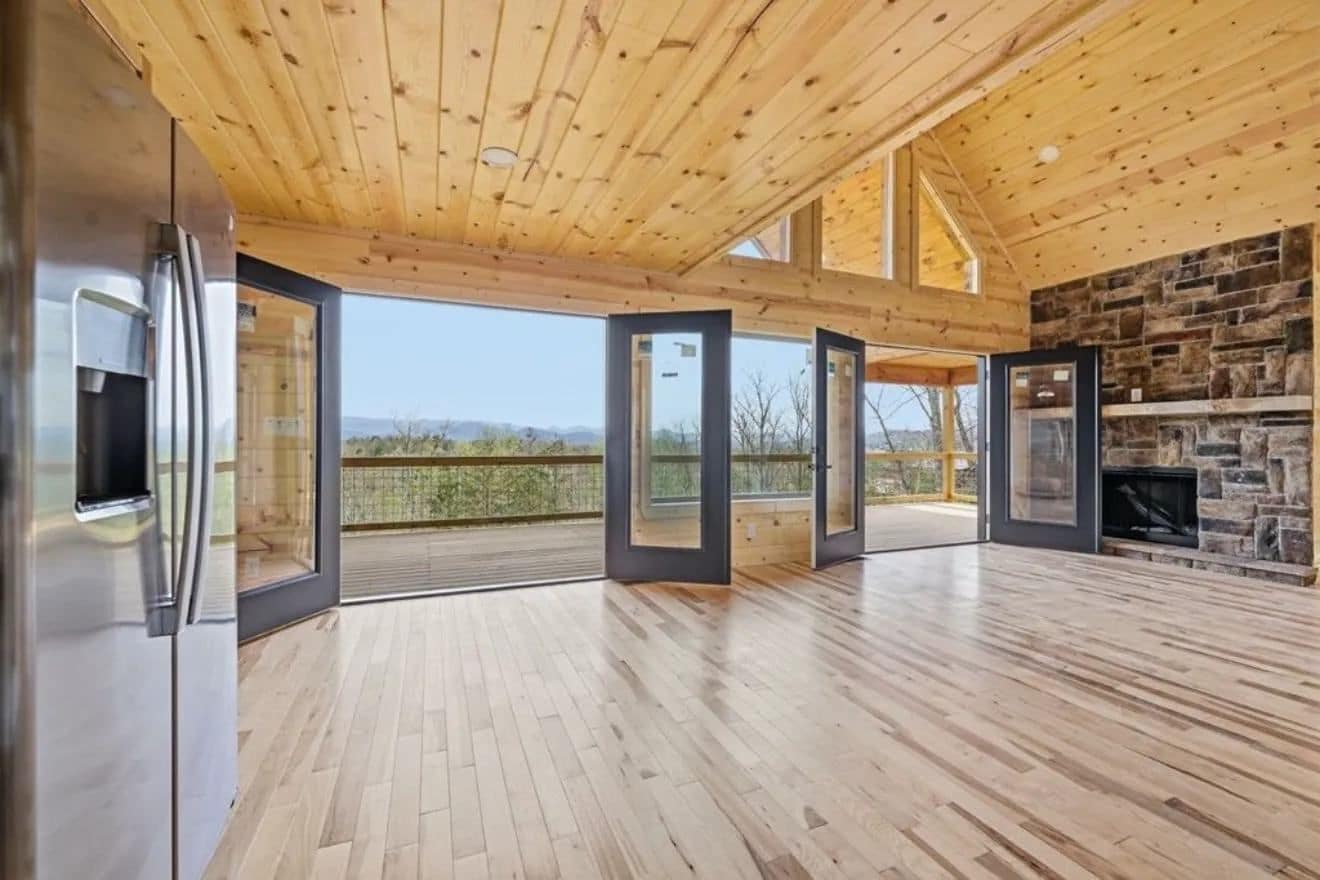 Open great room with a pine plank ceiling, a stone fireplace wall, a stainless refrigerator and black-framed glass doors opening to a deck