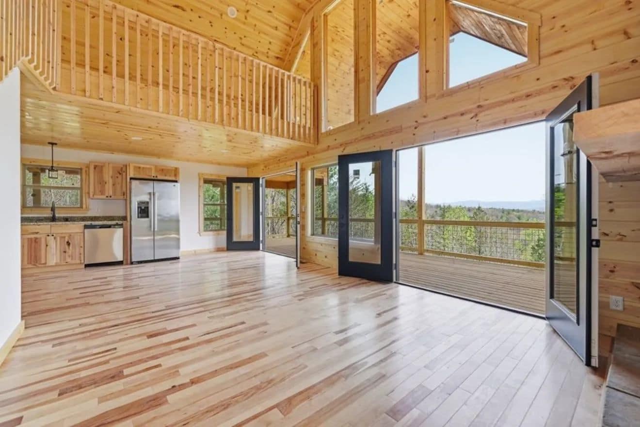 Great room with a loft railing above, a vaulted pine ceiling, a kitchen at the back and glass doors to a deck