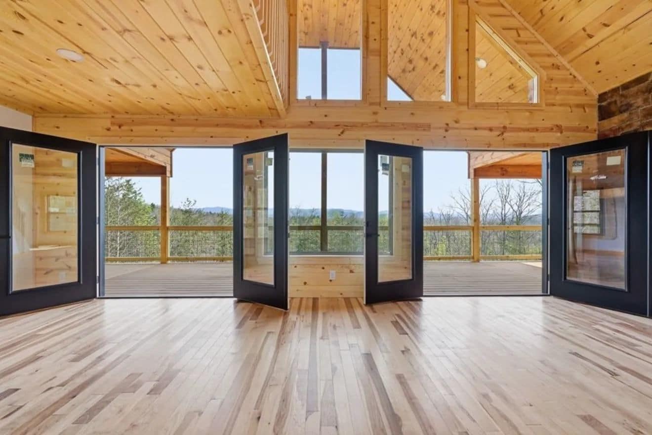 Open room with a high pine ceiling, hardwood floor and a wall of black-framed glass doors onto a deck