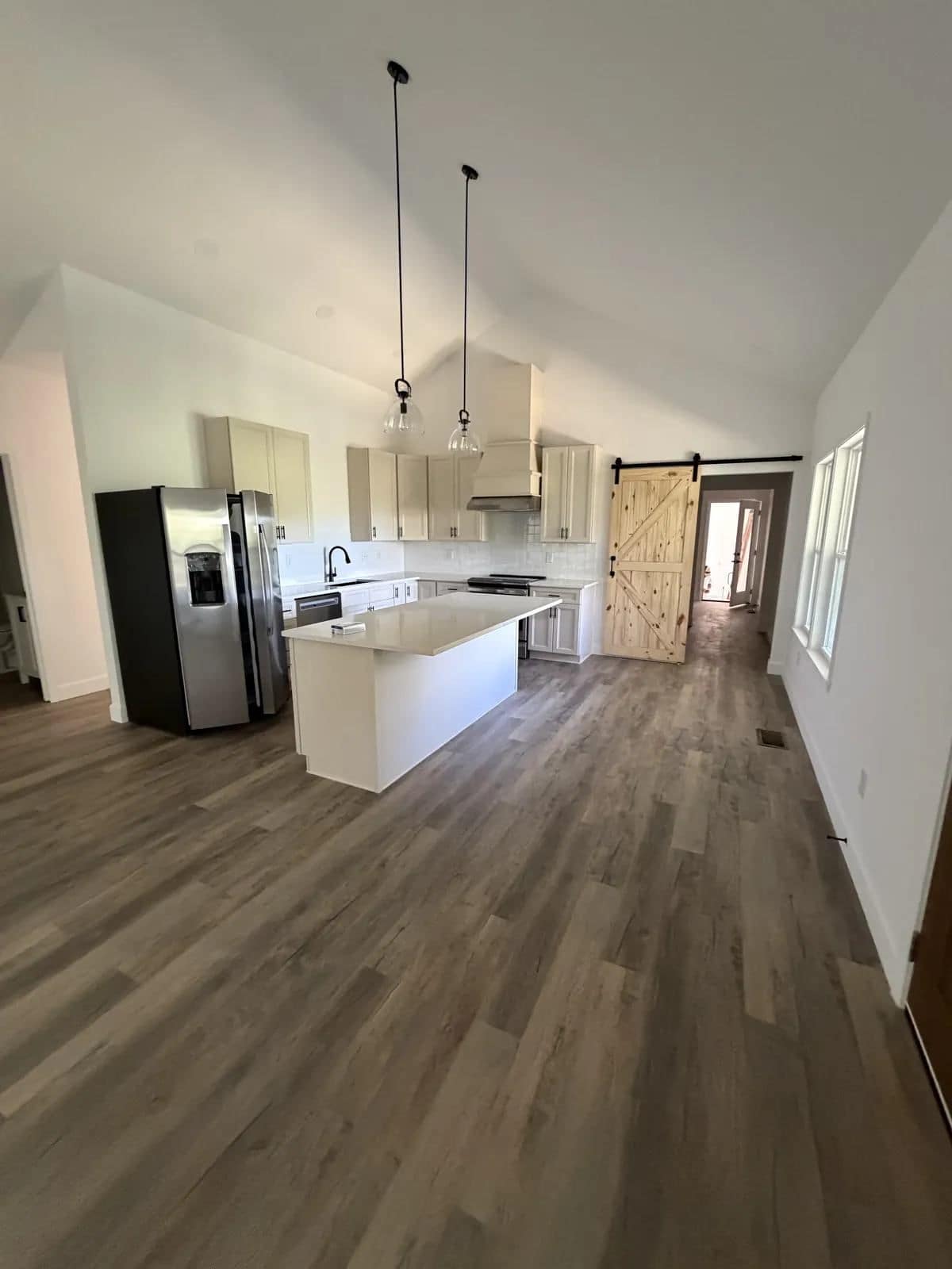 Kitchen with white cabinets, a large island, pendant lights, a stainless refrigerator and a barn door, over gray plank flooring