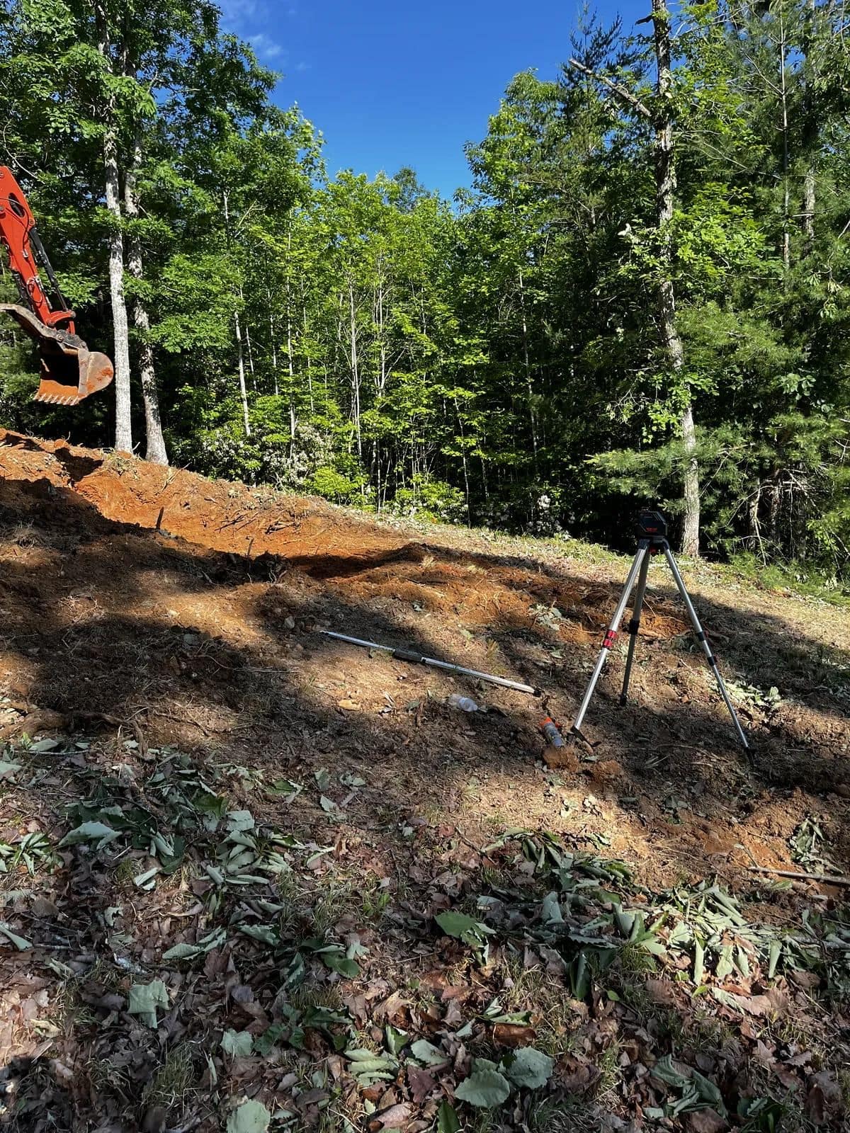 Cleared hillside with bare soil, a surveying tripod and an orange excavator at the edge